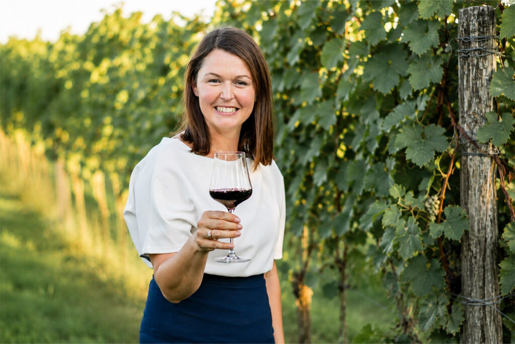 Eine Frau steht in einem Weinberg, lächelt und hält ein Glas Rotwein in der Hand, im Hintergrund sind grüne Weinreben zu sehen.